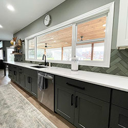 A long kitchen counter area with drawers, a dishwasher, and sink sits below a large kitchen window and white cabinetry